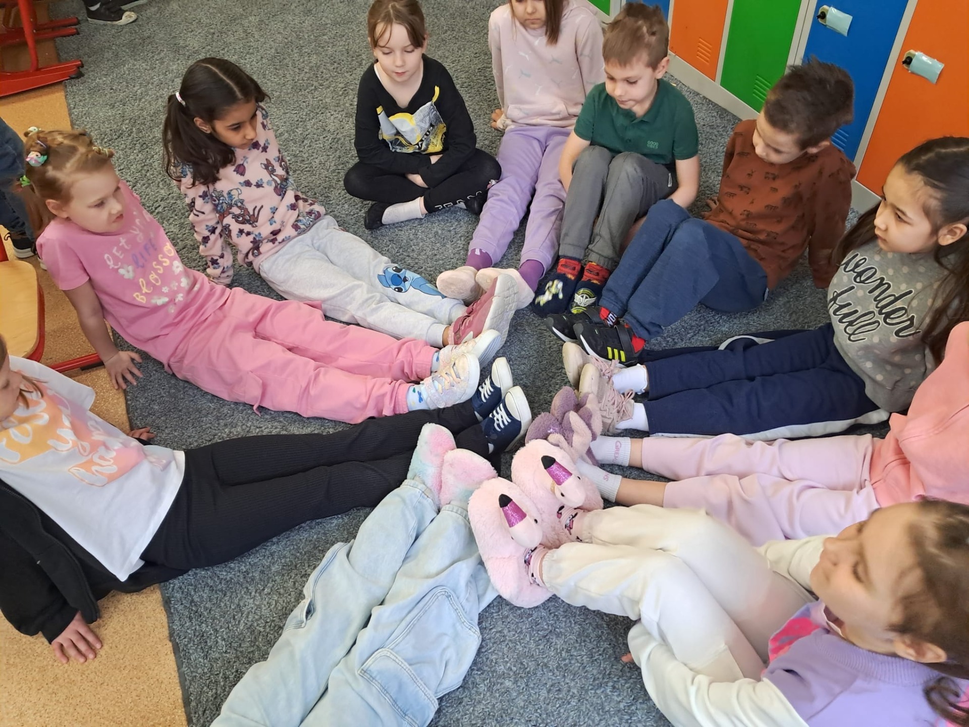 Uczniowie i uczennice klasy pierwszej siedzą w sali lekcyjnej na dywanie w kręgu. Dzieci mają wyprostowane nogi i stykają się kapciami. Każdy ma jakieś inne papcie pluszowe i kolorowe.