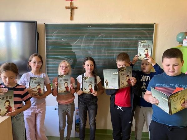  Dzieci w szkole stojące pod tablicą z uniesionymi książkami