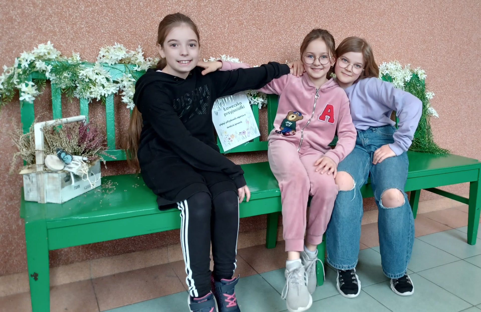 Trzy kolorowo ubrane dziewczynki siedzą na zielonej ławce przyjaźni obejmując się.