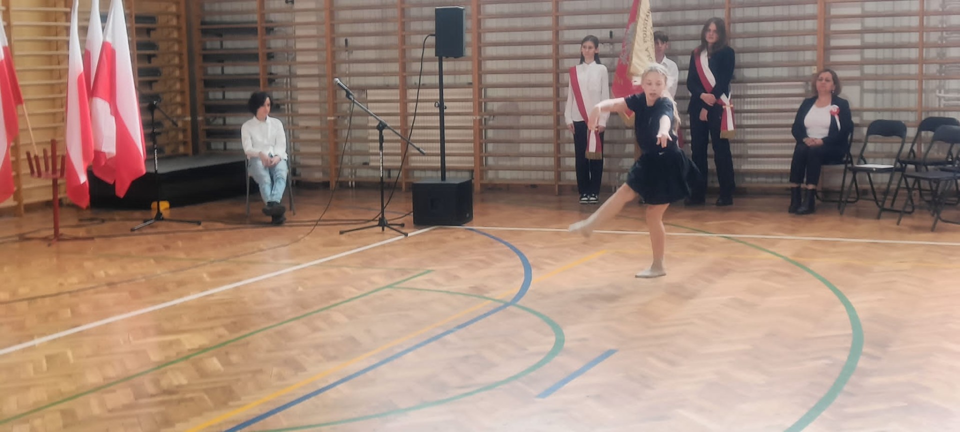 Uczennica tańczy  na sali gimnastycznej w tle Poczet Sztandarowy ze sztandarem SP1 Żory 