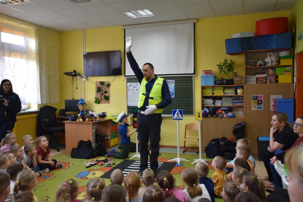 Policjant pokazuje przedszkolakom znaki ręcznego kierowania ruchem.