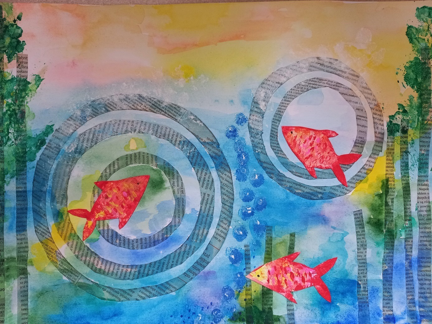 Praca malarska przedstawia trzy czerwone rybki w kręgach i wodorostach wykonanych z gazety na niebiesko-błękitnym tle