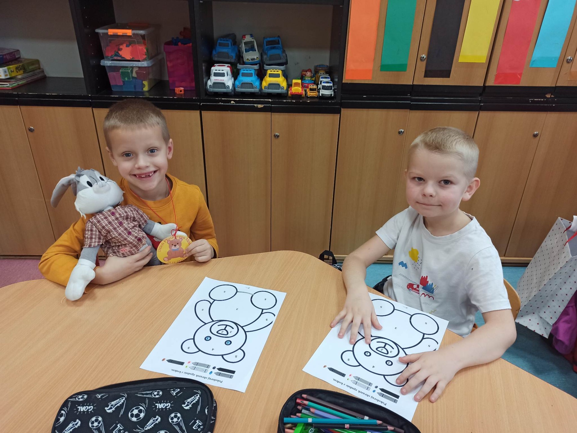 Dzieci siedzą przy stolikach w sali przedszkolnej i kolorują rysunki z misiem