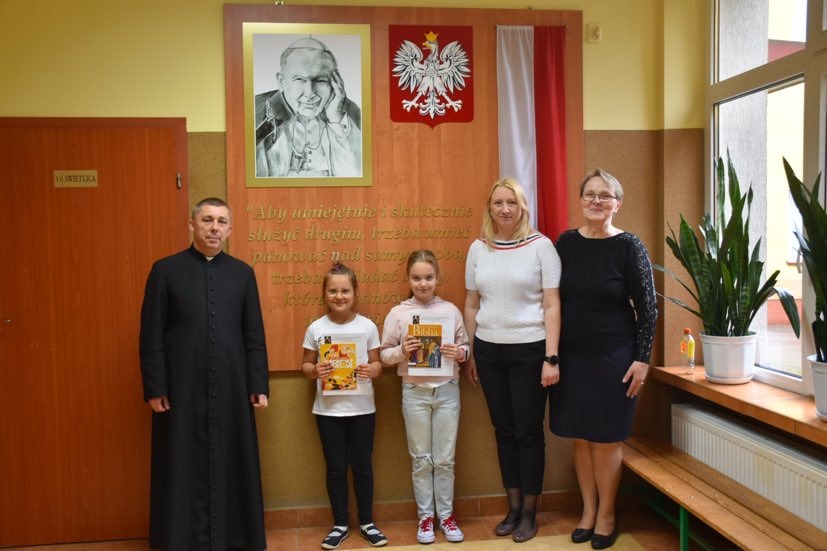Dwie uczennice wraz z księdzem, dyrektor i wicedyrektor szkoły przed tablicą pamiątkową ze zdjęciem Jana Pawła II