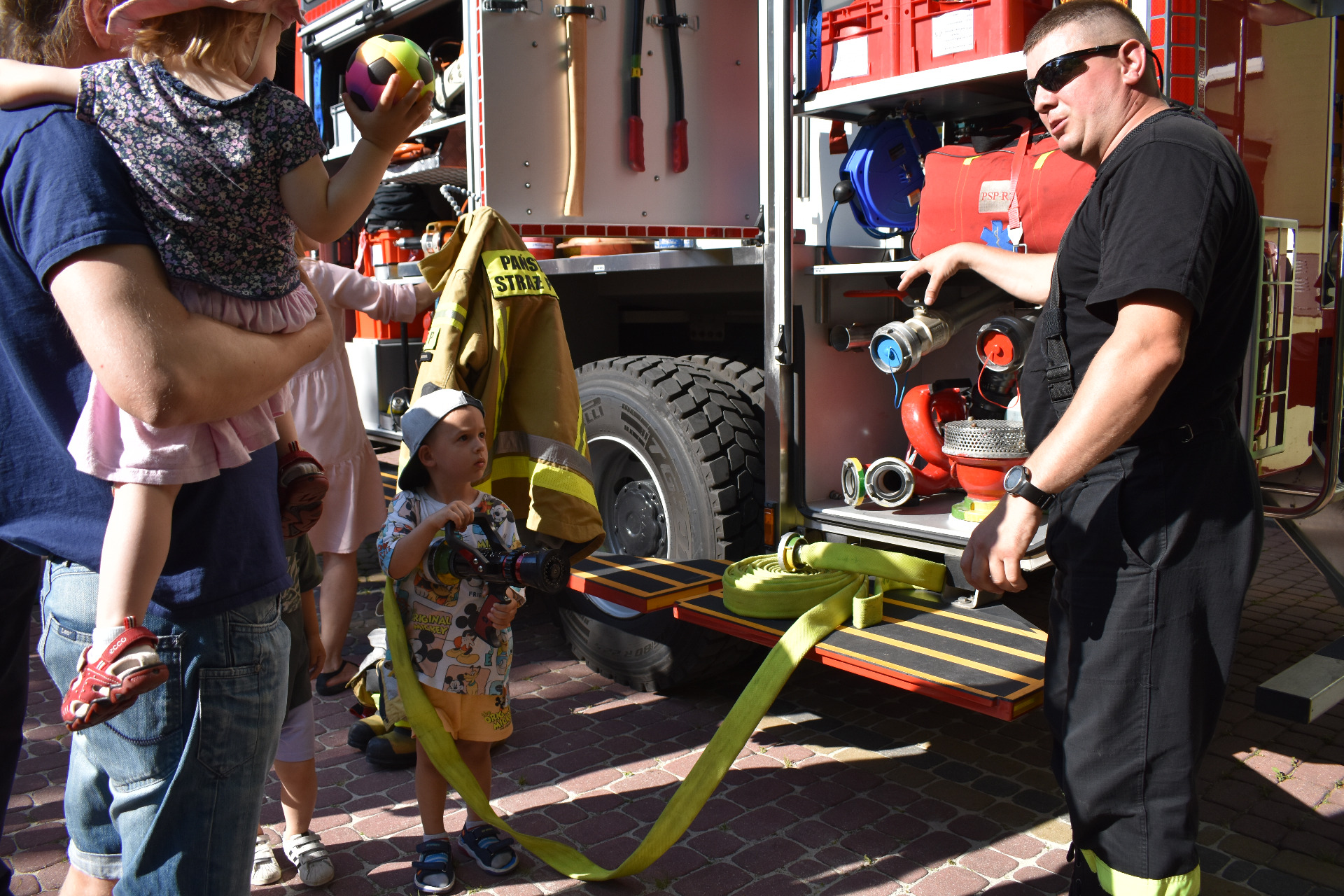 Chłopiec przy wozie strażackim trzymający węża. Tata z synkiem na rączkach podczas rozmowy z strażakiem o sprzęcie strażackim.