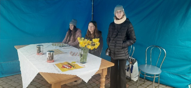 3 dziewczynki pod niebieskim namiotem, dwie dziewczynki siedzą przy stoliku, jedna dziewczynka stoi. Na stoliku stoją dwie puszki, stoi wazon z kwiatami oraz plakat.