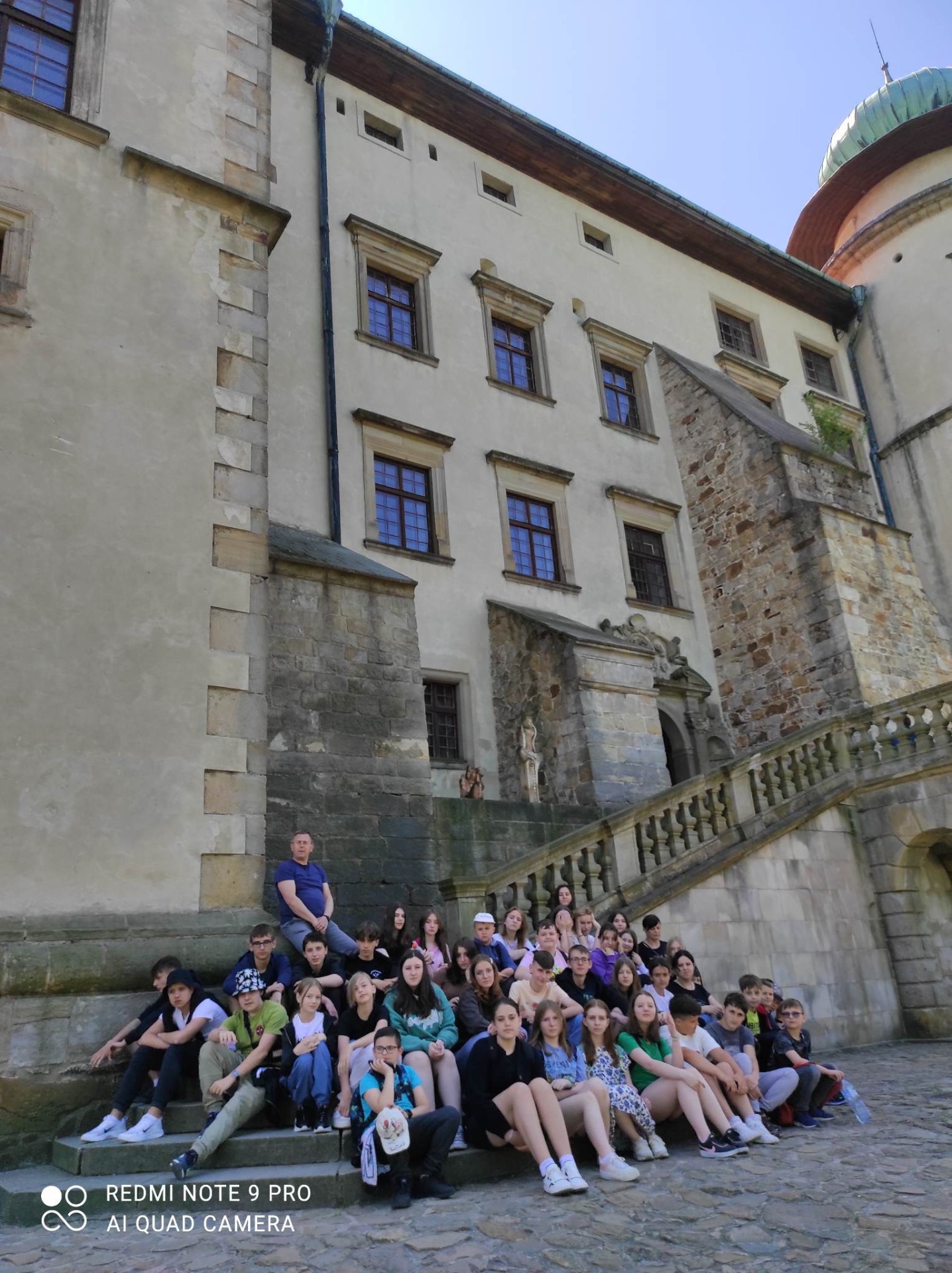 Zdjęcie przedstawia grupę uczniów i nauczycieli siedzących na schodach na zewnątrz zamku w Wiśniczu.