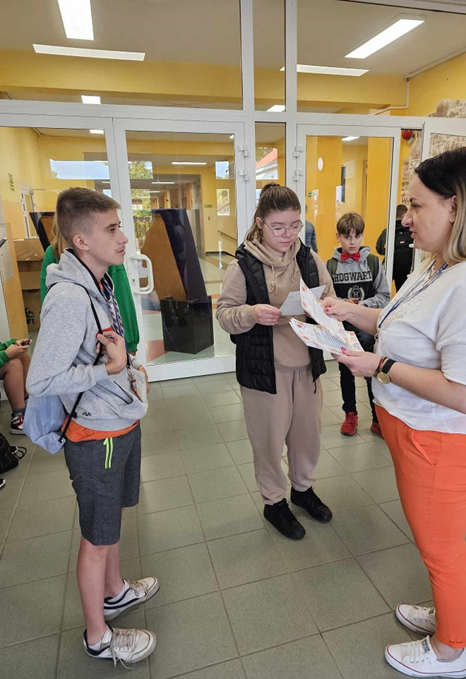 Pani pedagog, Magdalena Kacprzak rozdaje uczniom ulotki na szkolnym korytarzu.