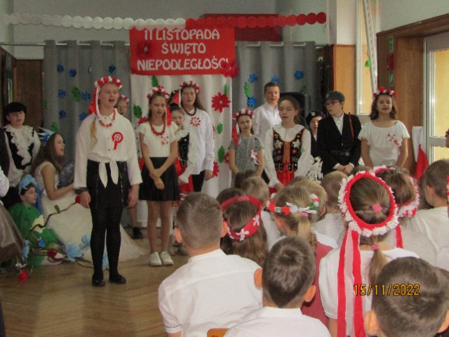 14 uczniów ubranych w czarno-białe stroje śpiewa piosenkę. Nad nimi wisi czerwony plakat z białym napisem 11 listopada Święto Niepodległości.

