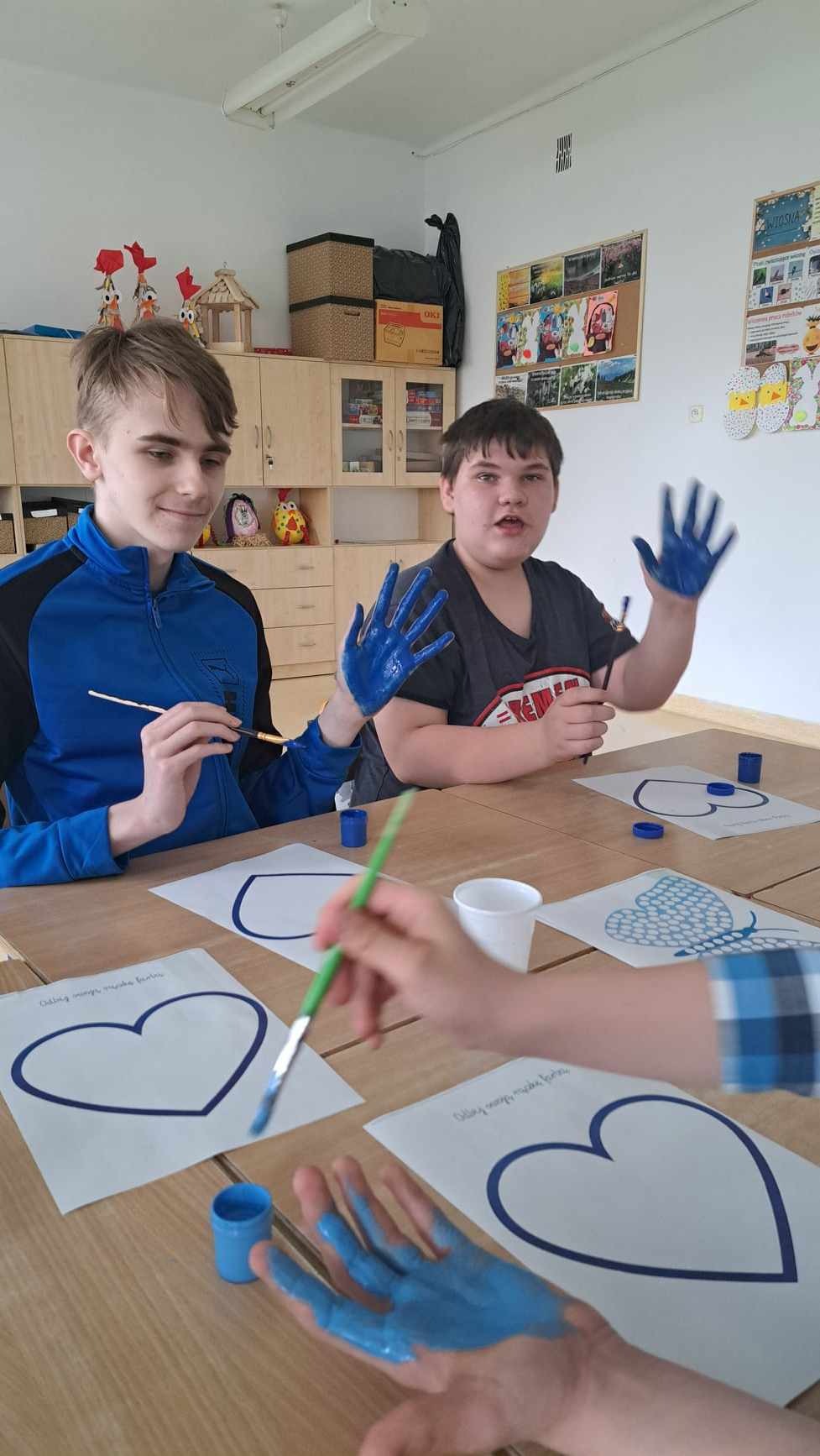 Uczniowie siedzą przy stoliku i wnętrze swojej dłoni malują niebieską farbą